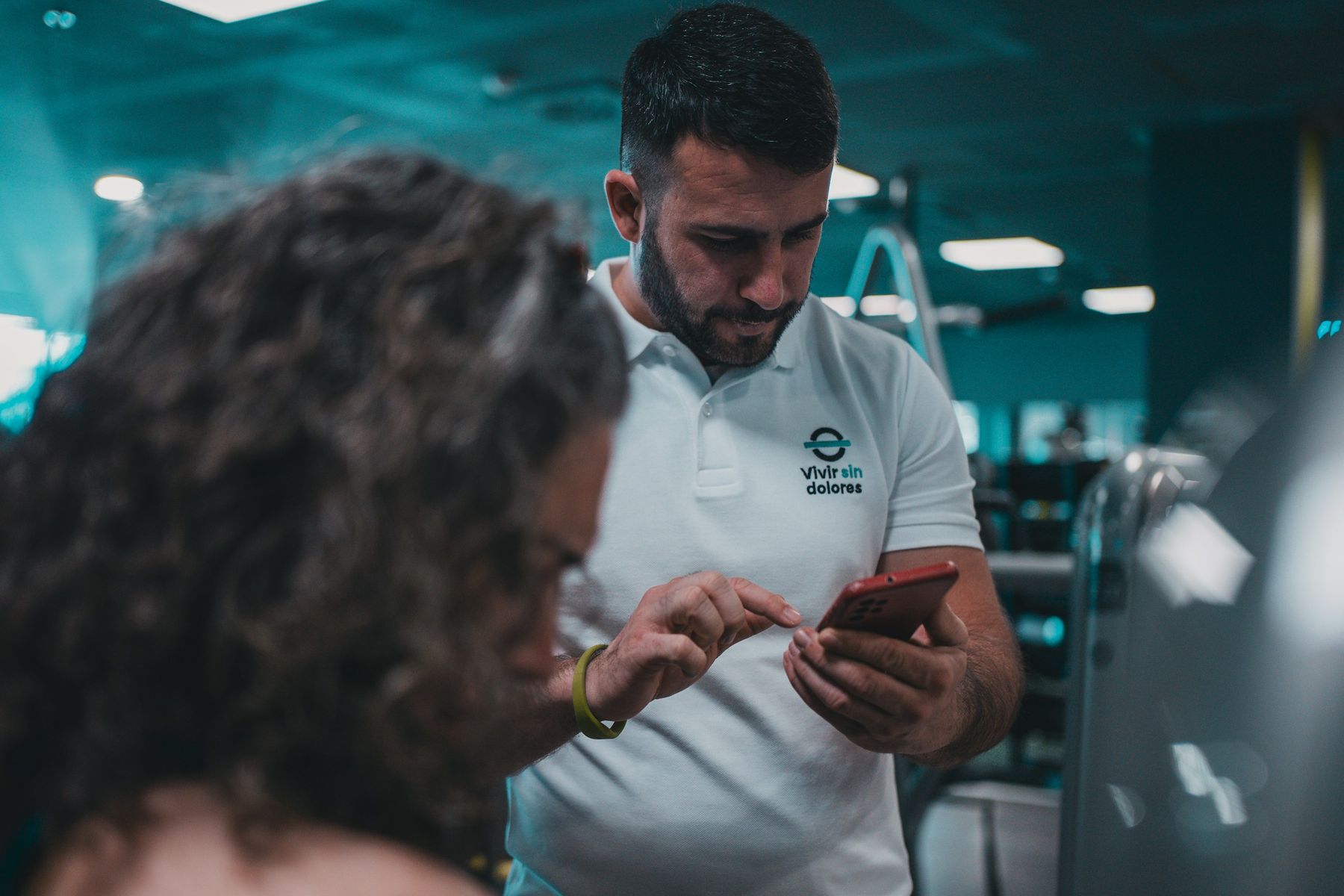 A staff member checks something on a phone with a member, a quiet one-to-one conversation on the gym floor.