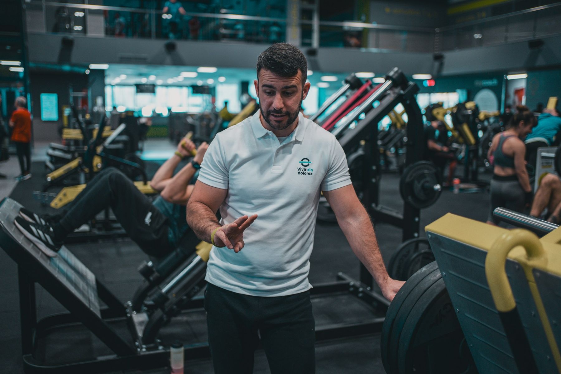 A coach in a white polo talks a member through the leg press on a busy gym floor while others train in the background.