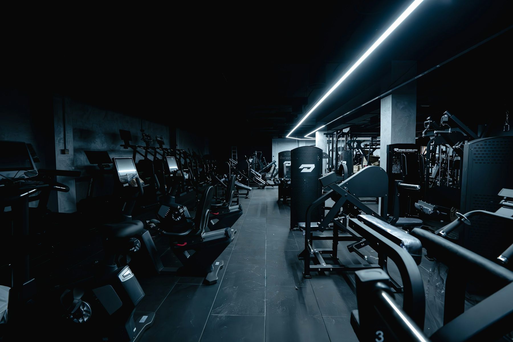 An empty gym after closing, with rows of cardio and strength machines under a single strip of light.