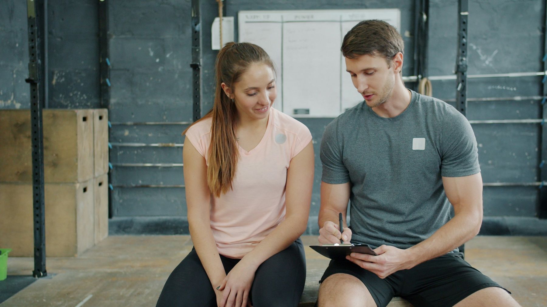 A trainer sits beside a new member in a functional-training gym, writing up her plan on a clipboard as she looks on.