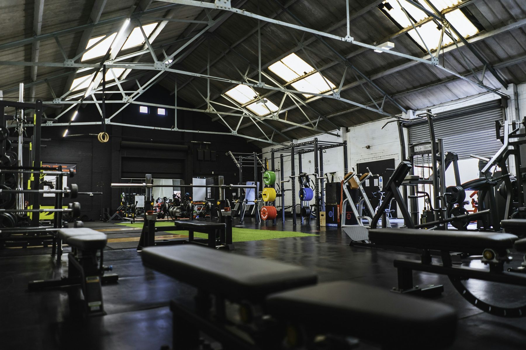A wide view of an independent warehouse gym with skylights, racks, benches and a turf lane.