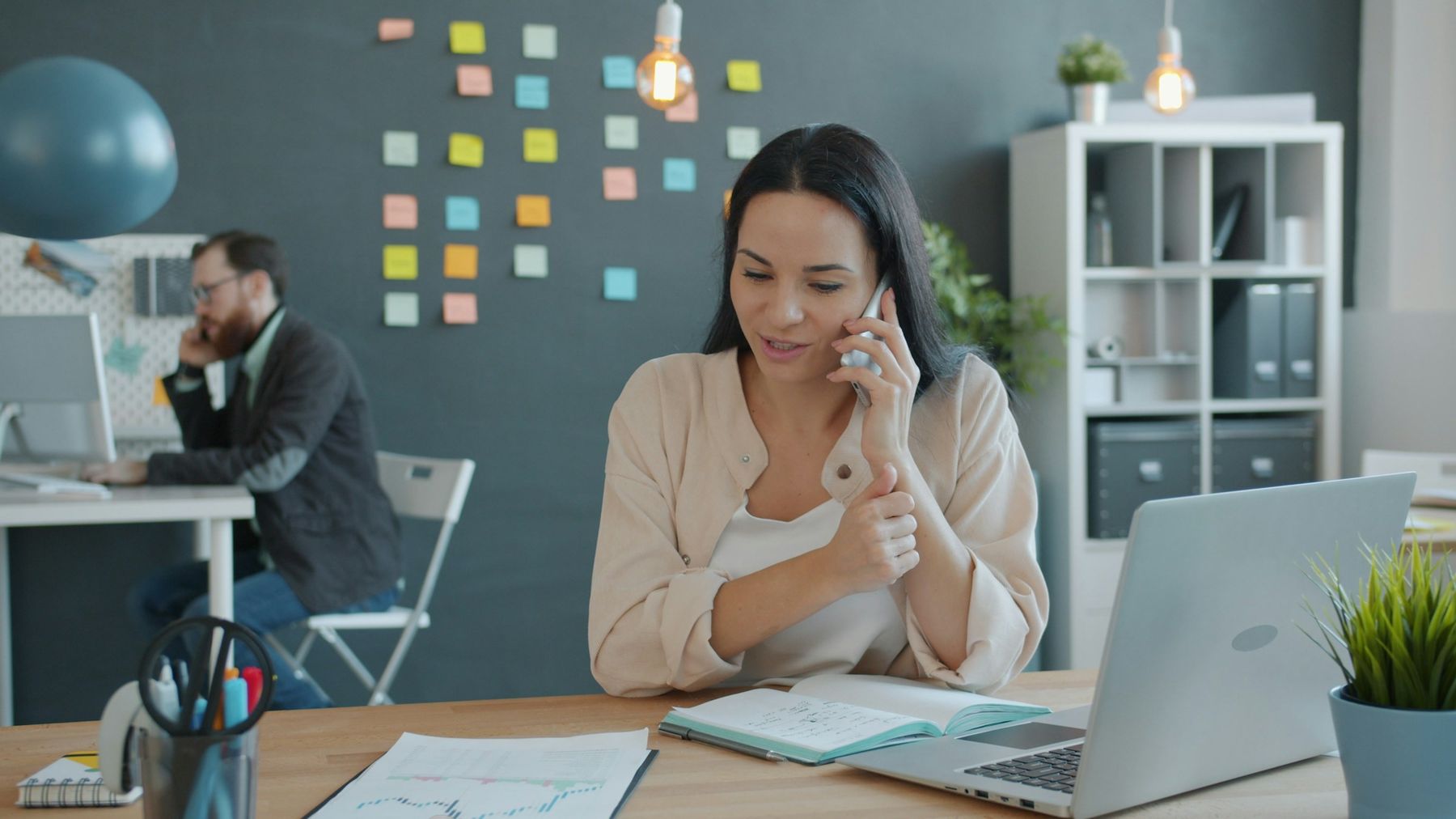 A manager makes a phone call at her desk in a small, bright back office, with a notebook open in front of her.
